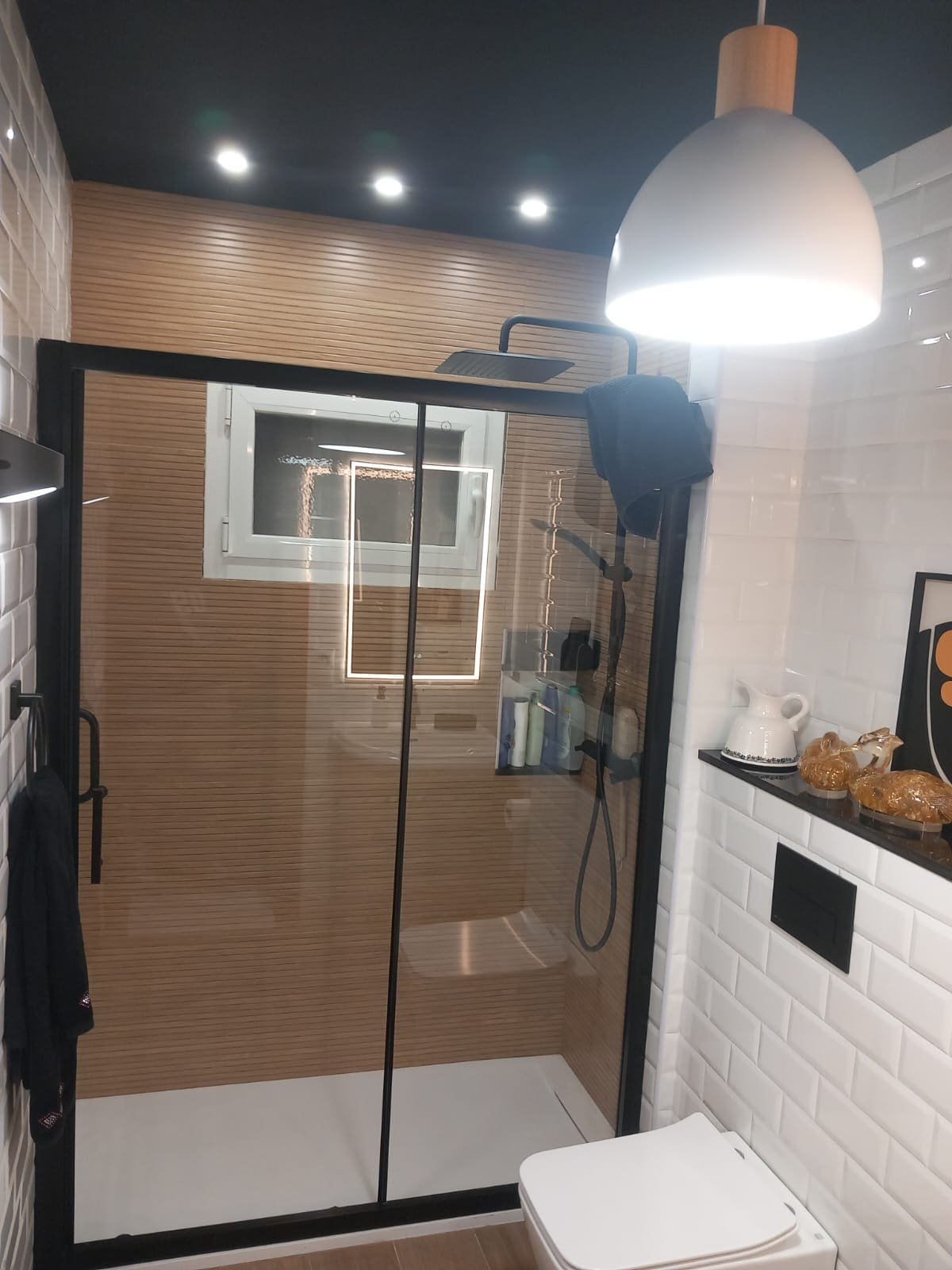 Baño con alicatado efecto madera, mampara negra y lámpara colgante sobre la ducha