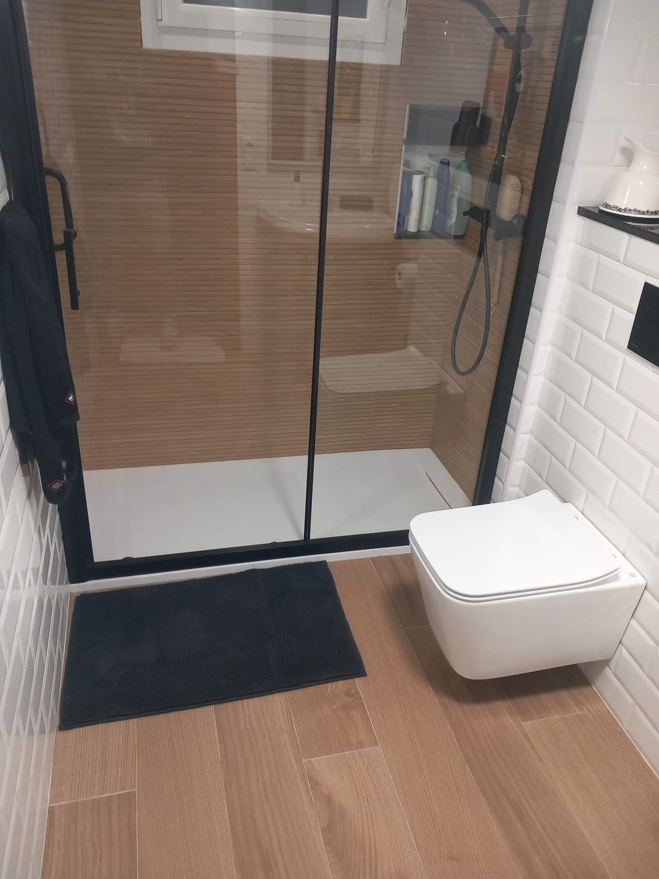Ducha con mampara de perfilería negra, azulejo metro blanco e inodoro suspendido